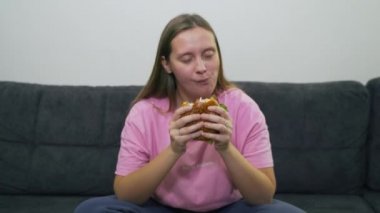 Mavi gözlü ve pembe tişörtlü tombul genç bir kadın gri koltukta oturuyor ve büyük bir zevkle koca bir hamburger yiyor. Kolesterol, abur cubur, alkol, sağlıksızlık, oburluk yağı