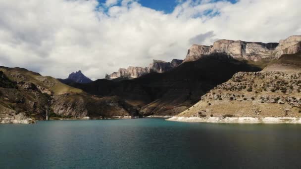 Luchtfoto van het azuurblauwe bergmeer Gizhgit, Kaukasus. Blauw water schijnt in het zonlicht. Hoge majestueuze bergketen is zichtbaar. Scherpe pieken van pieken gloeien tegen de bewolkte lucht. Drone videoshooting — Stockvideo