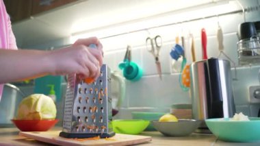 Kadın yemek pişirmek için mutfaktaki gümüş metal rende turuncu havuçları öğütüyor. Arka planda sebzeli renkli tabaklar var. Sağlıklı yiyecekler, sebzeler, evde yemek pişirmek.