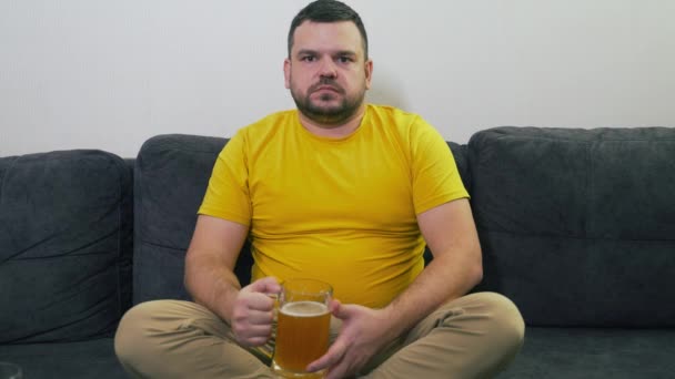 Homem de camiseta amarela está sentado no sofá cinza em casa em frente à TV e bebendo cerveja em grandes goles de caneca de vidro. Ele está tenso e atento. Relaxar em casa, ver televisão. Freelance. Álcool — Vídeo de Stock