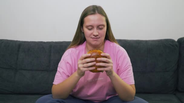 Mujer joven regordeta con ojos azules y camiseta rosa está sentado en el sofá gris y comer hamburguesa grande y gorda con gran placer. Colesterol, comida chatarra, alcohol, malestar, grasa de gula — Vídeos de Stock