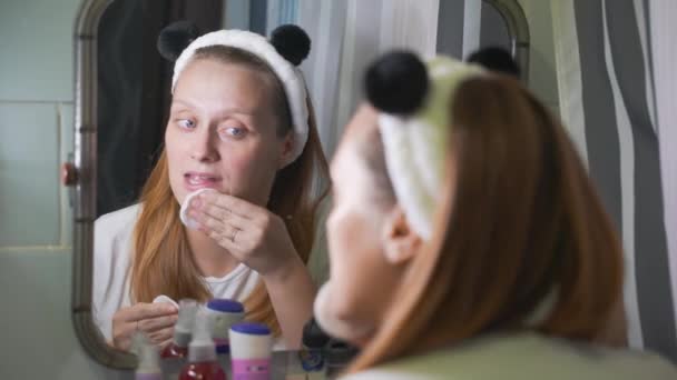 Jonge vrouw kijkt naar zichzelf in de badkamer spiegel en veegt de roodheid op haar gezicht met sponzen met lotion. Er zijn roodheid en puistjes op het gezicht. Gezondheid, cosmetologie, huidbehandeling — Stockvideo