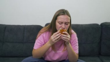 Mavi gözlü tombul genç kadın gri koltukta oturuyor ve büyük bir hamburger yiyor ve büyük cam bardaktan bira içiyor. Çöreği büyük lokmalarla ısırıyor. Kolesterol, abur cubur, alkol, sağlıksız