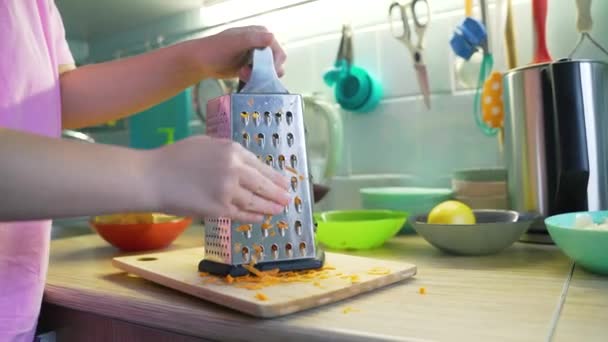 Frau zerkleinert orangefarbene Karotten auf silberner Metallreibe in der Küche auf blauem Brett zum Kochen. Im Hintergrund stehen bunte Teller mit Gemüse. Gesundes Essen, Gemüse, Kochen zu Hause — Stockvideo