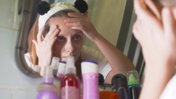 Jeune femme caucasienne se regarde dans le miroir de la salle de bain et examine la peau de son visage et se fâche. Il y a des rougeurs et des boutons sur le visage. La fille est bouleversée. Santé, cosmétologie — Video