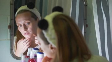 Genç kadın banyo aynasında kendine bakar, yüzünün derisini inceler, sivilceyi bulur ve sıkar. Yüzünde kızarıklık ve sivilceler var. Sağlık, kozmetoloji, cilt tedavisi