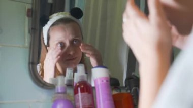 Genç beyaz kadın banyo aynasında kendine bakıyor ve yüzünün derisini inceliyor ve üzülüyor. Yüzünde kızarıklık ve sivilceler var. Kız çok üzgün. Sağlık, kozmetoloji