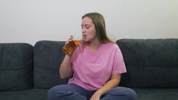 Junge kaukasische Frau in rosa T-Shirt sitzt zu Hause auf grauem Sofa und trinkt Bier aus durchsichtigem Glas. Sie lacht und lächelt. Bierwerbung. Schlechte Angewohnheit, Alkohol, Alkoholismus, ungesunde Ernährung — Stockvideo