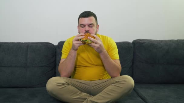 Giovane grassoccio con gli occhi marroni è seduto sul divano grigio e mangiare hamburger grande grasso. Morde il panino a pezzetti. Sete e fame. Colesterolo, dieta malsana, malessere, grasso di gola — Video Stock