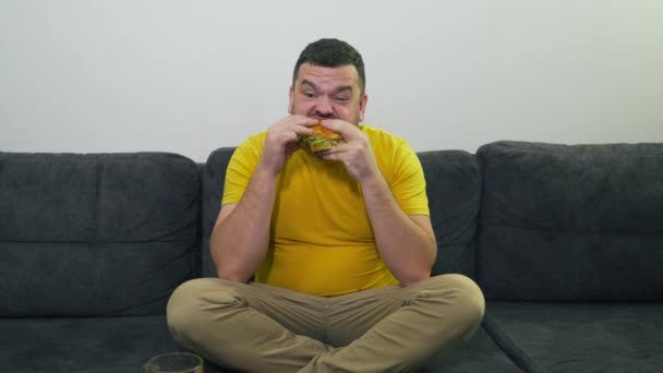 Een dikke jongeman met bruine ogen zit op een grijze bank en eet een dikke hamburger. Hij bijt het broodje in grote stukken. Dorst en honger. Cholesterol, ongezond dieet, ongezond, vet met overvloed — Stockvideo