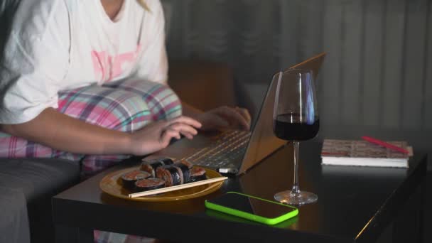Žena v pyžamu a brýlích sedí na gauči a doma pracuje na laptopu. Pozdě večer, tma. Na stole je porce jídla a sklenice vína. Práce doma, samovazba, na volné noze — Stock video