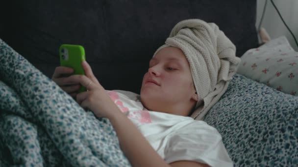 Young woman with towel on her head is lying in a straightened bed under blanket in pajamas and looking at the smartphone screen. Shes reading something on her phone and typing. Study, work at home — Stockvideo