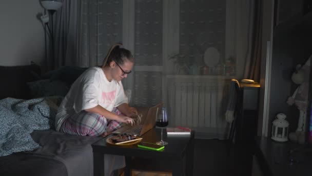 Mujer en pijama y gafas está sentado en el sofá y trabajando en el ordenador portátil en casa. Tarde en la noche, oscuro. Hay una porción de comida y una copa de vino sobre la mesa. Trabajo en casa, autoaislamiento, freelance — Vídeos de Stock