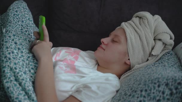 Young woman with towel on her head is lying in a straightened bed under blanket in pajamas and looking at the smartphone screen. Shes reading something on her phone and typing. Study, work at home — Vídeo de Stock