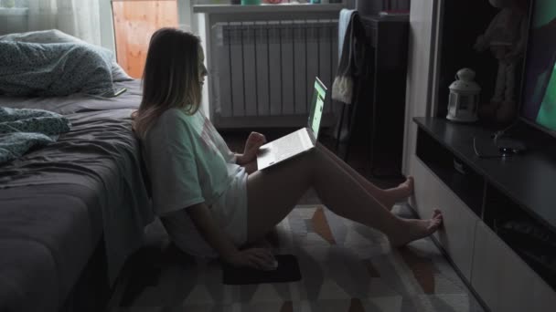 Молода жінка сидить на підлозі на килимі перед телевізором і працює на ноутбуці рано вранці. Вона спирається на диван, розкладений. Робота вдома, пандемія, бізнес, дистанція — стокове відео