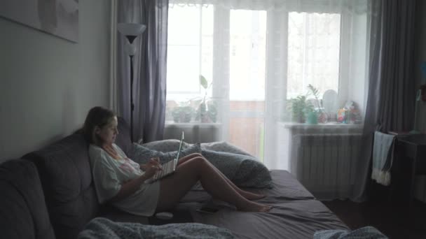 La mujer está sentada en el sofá enderezado y trabajando en el ordenador portátil en la mañana nublada temprano en casa. Ella mira la pantalla de la computadora y escribe algo en el teclado. Trabajo en casa, estudio, pandemia — Vídeos de Stock