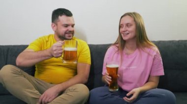 Genç çift gri kanepede oturuyor ve televizyon izliyorlar. Temiz bardakları tokuşturup bira içiyorlar. Keyfin yerinde. Onlar şaşırmış, gülüyor ve gülümsüyor. Rahatlamak, aşık olmak, televizyon izlemek.