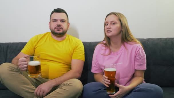 Молода пара сидить на сірому дивані, дивиться телевізор і п'є пиво з прозорих окулярів. Гарний настрій. Вони здивовані, сміються і посміхаються. Розслабляючись, закохуючись, дивиться телевізор — стокове відео