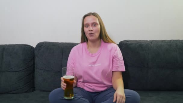 핑크 티셔츠를 입은 젊은 여성 이 집에 있는 회색 소파에 앉아 투명 한 유리 에서 TV 를 보고 맥주를 마시고 있다. 그녀는 놀라고 웃는다. 나쁜 습관, 술, 알코올 중독, 우울증 — 비디오