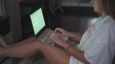 Genç bir kadın TV 'nin karşısında halıda oturuyor ve bulutlu bir sabahta dizüstü bilgisayarla çalışıyor. Kanepeye uzanmış yatıyor. Evde çalışmak, salgın, iş, mesafe