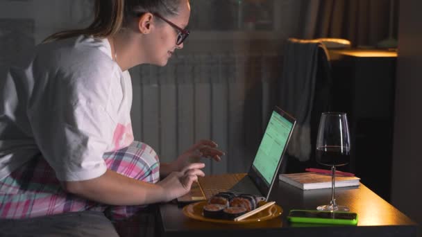 Woman in pajamas and glasses is sitting on sofa and working on laptop at home. Late evening, dark. There is portion of food and glass of wine on the table. Work at home, self-isolation, freelance — Stock Video