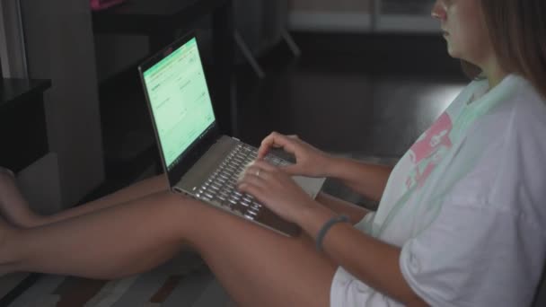 Young woman is sitting on the floor on carpet in front of the TV and working on laptop in the early cloudy morning. She leans back on the sofa spread out. Work at home, pandemic, business, distance — Stockvideo