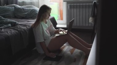 Genç bir kadın TV 'nin karşısında halıda oturuyor ve bulutlu bir sabahta dizüstü bilgisayarla çalışıyor. Kanepeye uzanmış yatıyor. Evde çalışmak, salgın, iş, mesafe