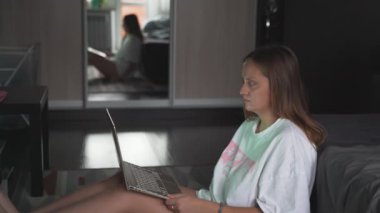 Genç bir kadın TV 'nin karşısında halıda oturuyor ve bulutlu bir sabahta dizüstü bilgisayarla çalışıyor. Kanepeye uzanmış yatıyor. Evde çalışmak, salgın, iş, mesafe