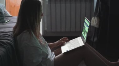 Genç bir kadın TV 'nin karşısında halıda oturuyor ve bulutlu bir sabahta dizüstü bilgisayarla çalışıyor. Kanepeye uzanmış yatıyor. Evde çalışmak, salgın, iş, mesafe