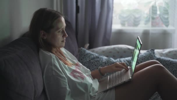 La mujer está sentada en el sofá enderezado y trabajando en el ordenador portátil en la mañana nublada temprano en casa. Ella mira la pantalla de la computadora y escribe algo en el teclado. Trabajo en casa, estudio, pandemia — Vídeos de Stock