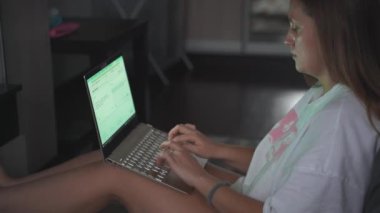 Genç bir kadın TV 'nin karşısında halıda oturuyor ve bulutlu bir sabahta dizüstü bilgisayarla çalışıyor. Kanepeye uzanmış yatıyor. Evde çalışmak, salgın, iş, mesafe