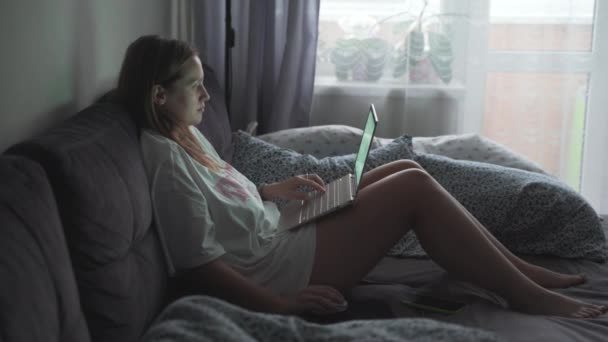 Woman is sitting on straightened sofa and working on laptop in the early cloudy morning at home. She looks at the computer screen and types something on the keyboard. Work at home, study, pandemic — 图库视频影像
