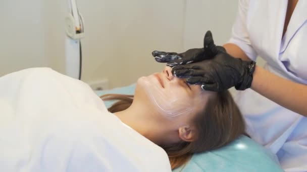 Cosmetologist applicerar vit mask på ljus hud av kvinnor ansikte i cirkulära rörelser före procedurer. Rengöring av orenheter. Doktorn har handskar på händerna. Hudrengöring, aknebehandling — Stockvideo