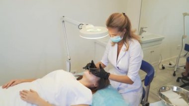 Kozmetik uzmanı, prosedürlerden önce kadınların güzel ciltlerine dairesel hareketlerle maske takıyor. Deriyi kirlilikten arındırıyor. Doktorun elinde eldivenler var. Deri temizleme, sivilce tedavisi