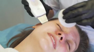 Kozmetik uzmanı deriyi ultrasonik temizlik cihazıyla temizliyor. Eldivenli doktorlar müşterinin yüzünü metal plakayla kaplıyor. Yenilenme prosedürü, sivilce ve sivilce tedavisi