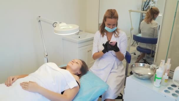 Cosmetologist in white coat is sitting in office and typing text on smartphone. Black gloves on his hands, protective mask on his face. Patient is lying in front of her. Break, online communication — Wideo stockowe