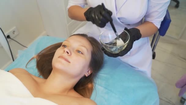 Kosmetolog menerapkan topeng untuk kulit wanita wajah untuk tujuan terapi. Pasien berbaring di sofa dan zat transparan tebal diterapkan untuk menghadapi kuas. Rejuvenasi, perawatan jerawat — Stok Video