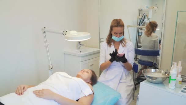 Cosmetologist in white coat is sitting in office and typing text on smartphone. Black gloves on his hands, protective mask on his face. Patient is lying in front of her. Break, online communication — 图库视频影像