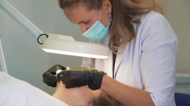 Doktor, kadının yüzündeki sivilceleri çıkartıyor. Siyah eldivenli eller steril süngerleri tutar ve açık tenli sivilceleri temizler. Lamba ve büyüteç, yüzü aydınlatır. Cilt temizleme işlemi