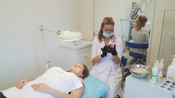 Cosmetologist in white coat is sitting in office and typing text on smartphone. Black gloves on his hands, protective mask on his face. Patient is lying in front of her. Break, online communication — Stock videók