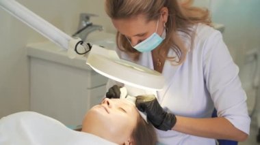 Doktor, kadının yüzündeki sivilceleri çıkartıyor. Siyah eldivenli eller steril süngerleri tutar ve açık tenli sivilceleri temizler. Lamba ve büyüteç, yüzü aydınlatır. Cilt temizleme işlemi