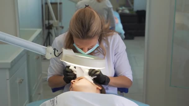 El doctor exprime espinillas en la cara de la mujer. Las manos en guantes negros sostienen esponjas estériles y eliminan la suciedad y las espinillas de la piel clara. Lámpara y lupa iluminan la cara. Procedimiento de limpieza de la piel — Vídeos de Stock