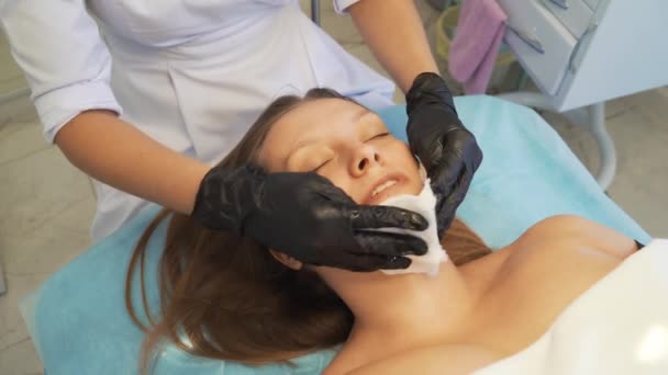 Косметолог вытирает лицо и шею молодой женщины из грязи влажными белыми губками. Она нежно трёт пациентам светлую кожу, чтобы очистить её от загрязнений. Косметология, лечение акне, акне, омоложение — стоковое видео