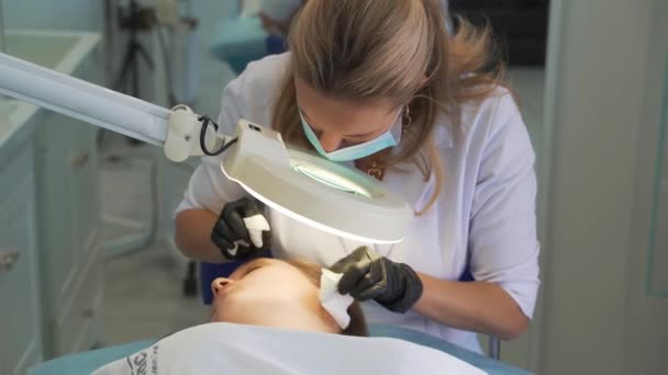 Doctor squeezes out pimples on face of woman. Hands in black gloves hold sterile sponges and remove dirt and pimples from fair skin. Lamp and magnifying glass illuminate face. Skin cleansing procedure — 图库视频影像