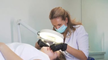 Doktor, kadının yüzündeki sivilceleri çıkartıyor. Siyah eldivenli eller steril süngerleri tutar ve açık tenli sivilceleri temizler. Lamba ve büyüteç, yüzü aydınlatır. Cilt temizleme işlemi