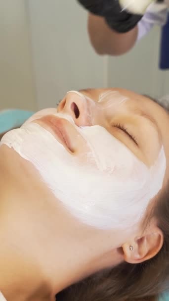 美容师为治疗目的将面罩涂在女性面部皮肤上。病人躺在沙发上，用厚厚的白色物质用刷子刷脸部。康复、痤疮治疗 — 图库视频影像