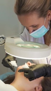 Doktor, kadının yüzündeki sivilceleri çıkartıyor. Siyah eldivenli eller steril süngerleri tutar ve açık tenli sivilceleri temizler. Lamba ve büyüteç, yüzü aydınlatır. Cilt temizleme işlemi