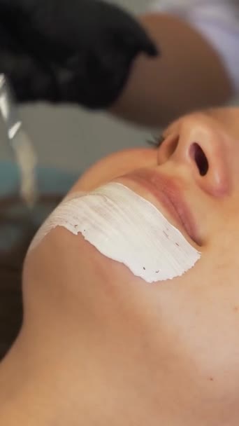 Cosmetologist applies mask to skin of womans face for therapeutic purposes. Patient is lying on couch and thick white substance is applied to face with brush. Rejuvenation, acne treatment — Wideo stockowe