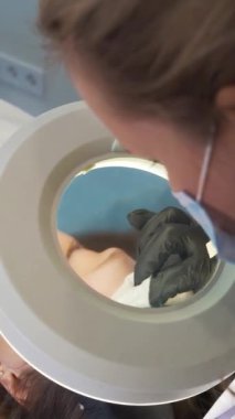 Doktor, kadının yüzündeki sivilceleri çıkartıyor. Siyah eldivenli eller steril süngerleri tutar ve açık tenli sivilceleri temizler. Lamba ve büyüteç, yüzü aydınlatır. Cilt temizleme işlemi