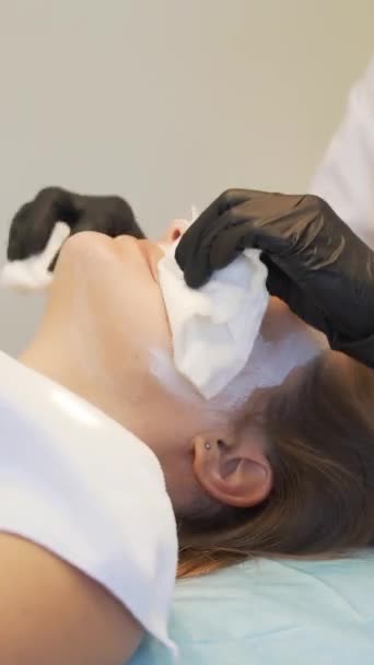 Cosmetologist tar bort vit mask från rättvis hud av unga kvinnor ansikte. Läkaren torkar tjockt vitt ämne från ansiktet med vita fuktiga svampar. Föryngring, vit lera, hudrengöring, aknebehandling — Stockvideo